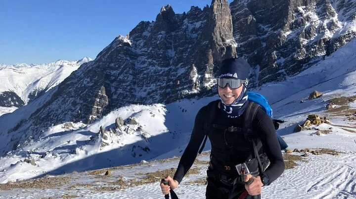 Skitourengeher lächelt in die Kamera während des Aufstiegs, im Hintergrund sieht man Berge | © Fabian Brormann