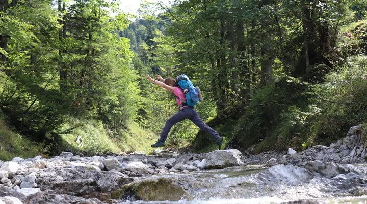Mädchen quert einen Bach und springt dabei über die Steine, die Hände zeigen in die Höhe. | © Waltraud Zopf